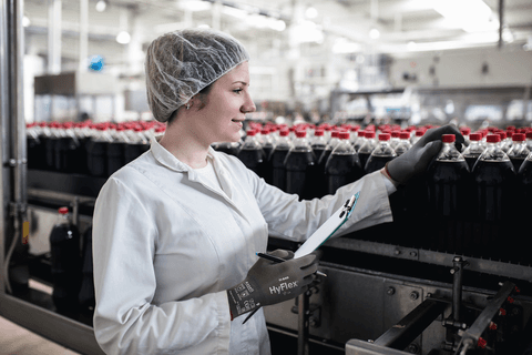 Worker in a food processing plant wearing food-approved HyFlex 11-840 gloves, inspecting bottles on a production line.